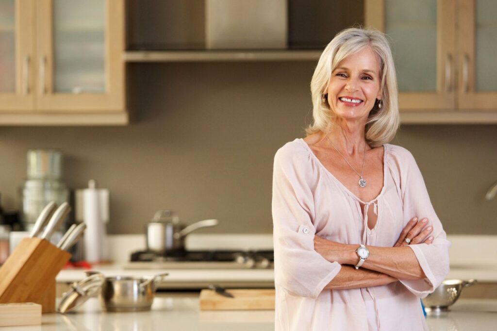 Older lady wearing a pink blouse standing in her kitchen with her arms folded and smiling.