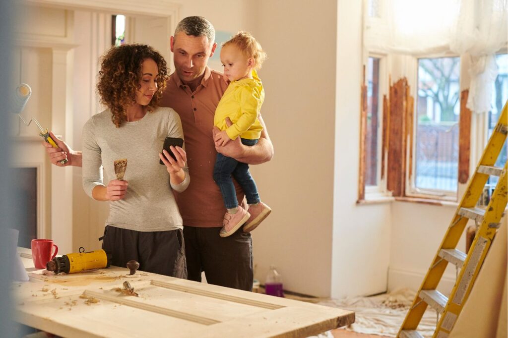 Young couple decorating their sitting room looking at a design on a mobile phone. The man is holding their toddler.